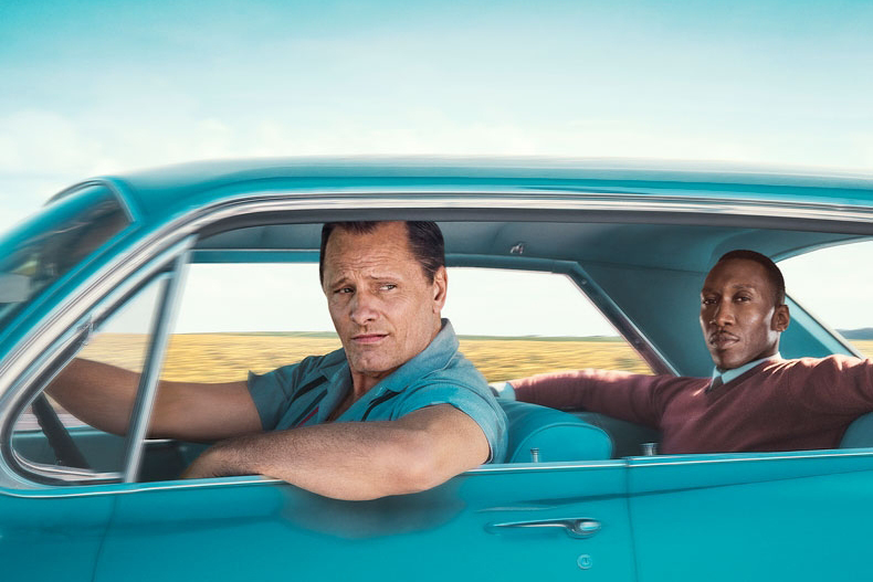 A white man and a black man sitting in the green car, and the window rolls down.